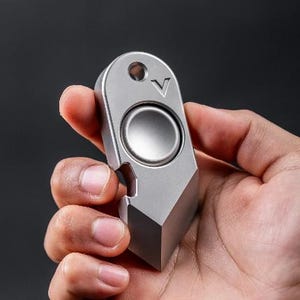 May include: A silver metal fidget spinner held in a hand. The spinner has a circular centre and a bottle opener feature. The top has a hole and a V-shaped marking. The object is a combination of a fidget toy and a tool.