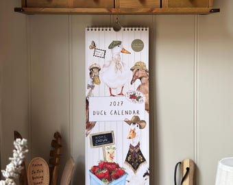 Puede incluir: Un calendario de pared para 2027 con ilustraciones de patos que llevan varios sombreros y accesorios. El calendario incluye un título central, un gráfico de una cesta azul llena de fresas rojas y un pequeño cartel con la etiqueta Home Grown Strawberries.