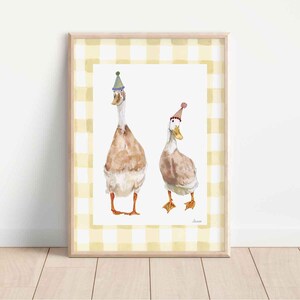 Op de afbeelding: Waterverfprint van twee ganzen met feesthoeden. De ganzen hebben bruine en witte veren en oranje poten. Het kunstwerk is ingelijst in een licht houten frame met een geel en wit geblokte rand. De achtergrond is wit.