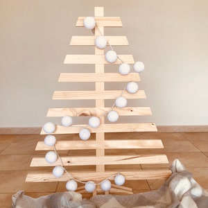 May include: A wooden Christmas tree with a white star on top and a string of white cotton ball lights wrapped around it. A white and gray plaid blanket is at the base of the tree.