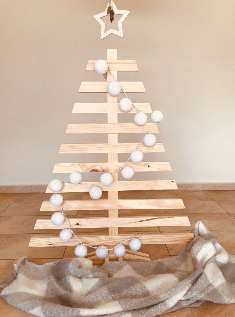 May include: A wooden Christmas tree with a white star on top and a string of white cotton ball lights wrapped around it. A white and gray plaid blanket is at the base of the tree.