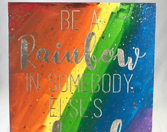 May include: A rainbow-colored background with silver lettering that reads "Be a rainbow in somebody else's cloud" and "Maya Angelou" at the bottom.