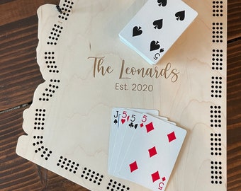May include: A wooden cribbage board in the shape of the state of Arizona. The board has a personalized inscription that reads "The Leonards Est. 2020". There are playing cards on the board, including a Jack of Spades, a 5 of Hearts, a 5 of Clubs, and a 5 of Diamonds.