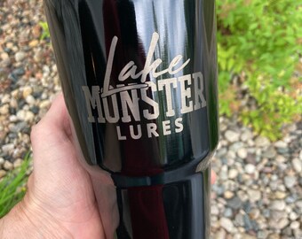 May include: Black stainless steel tumbler with a silver inscription that reads "Lake Monster Lures".