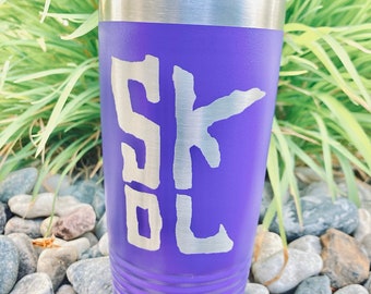 May include: Purple stainless steel tumbler with a white "SKOL" graphic. The tumbler has a lid and is sitting on a bed of small rocks.