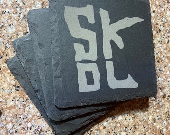 May include: A stack of six black slate coasters with the word "SKOL" written in white on the top coaster.