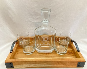 May include: A wooden serving tray with black metal handles, featuring a clear glass decanter and four glasses. Each glass and the decanter are etched with the letter "B".