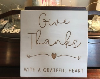 May include: White wooden sign with the text "Give Thanks with a Grateful Heart" in brown cursive lettering. A small heart is drawn between the words "Give" and "Thanks".