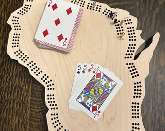 May include: A wooden game board shaped like the state of Wisconsin with a peg game and playing cards. The board has holes for pegs and the text "START" and "HOME" are visible. The cards are a five of diamonds, a five of clubs, a five of hearts, and a jack of diamonds.