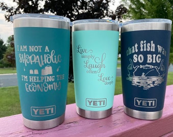 May include: Three stainless steel YETI tumblers with different engraved sayings. The first tumbler is turquoise blue with the saying "I am not a shopaholic, I'm helping the economy". The second tumbler is light blue with the saying "Live simply, laugh often, love deeply". The third tumbler is dark blue with the saying "That fish was so big".