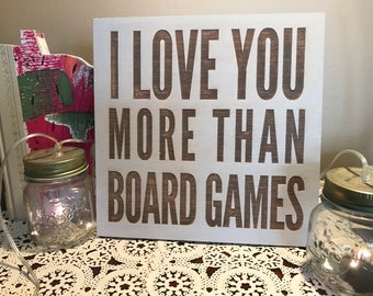 May include: A white wooden sign with the text "I love you more than board games" in brown lettering.