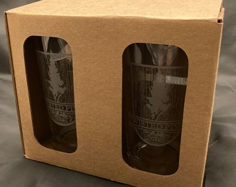 May include: Two clear glass pint glasses with a brown cardboard box. The glasses have an etched design of a pine tree and the words "Twisted Pine" and "Salem".