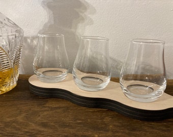 May include: Three clear glass whiskey glasses sit on a wooden coaster with a wavy edge. The coaster is black and white.