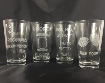 May include: Four clear glass drinking glasses with humorous phrases etched into the glass. The phrases are "Can't Recognize These My Tracks", "Smores Burger", "How Neat Is That? That's Pretty Neat", and "Tree Poop".