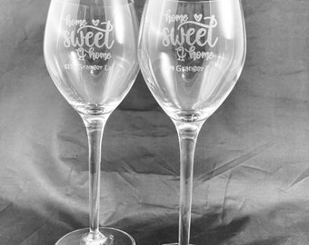 May include: Two clear glass wine glasses with the text "home sweet home" etched on the front. The glasses are on a black surface.