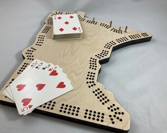 May include: A wooden game board shaped like the state of Minnesota with a black border and holes for pegs. A deck of playing cards is on the board, including a stack of 5 of diamonds and a hand of 5 of hearts, 5 of clubs, and Jack of spades.