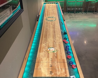 May include: A wooden shuffleboard table with a green felt surface and a duck logo. The table has blue LED lights around the perimeter and is set up for play with red and blue discs.