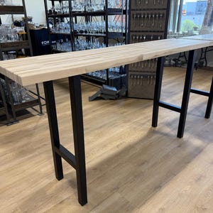 May include: A long, rectangular wooden table with a light-colored top and black metal legs. The table is supported by two sets of black legs, creating a sturdy base. The table is approximately 10 feet long.