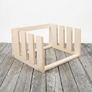 May include: A light-colored, unfinished wooden crate with vertical dividers. The crate is rectangular with an open top and is constructed from multiple pieces of wood. The crate sits on a weathered, gray wooden surface.