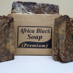 May include: Blocks of Africa Black Soap, with a cardboard box in the background. The box has the words "Africa Black Soap (Premium)" printed on it. The soap is dark brown and has a rough, textured appearance.