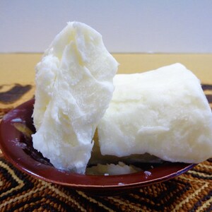 May include: Two large, solid, opaque white chunks of raw shea butter with a dense, waxy texture. The butter pieces are stacked and resting inside a small, round, dark reddish-brown bowl with a smooth, glossy finish and a slightly flared rim.