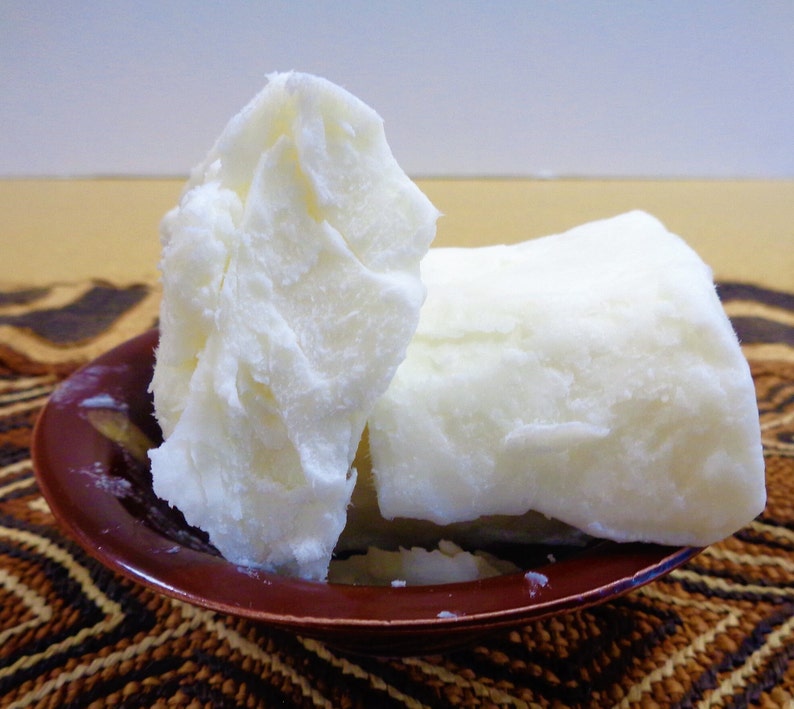 May include: Two large, solid, opaque white chunks of raw shea butter with a dense, waxy texture. The butter pieces are stacked and resting inside a small, round, dark reddish-brown bowl with a smooth, glossy finish and a slightly flared rim.