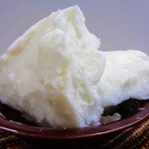 May include: A large, solid, off-white chunk of raw shea butter with a crumbly, uneven texture. The butter is piled high and rests inside a small, round, dark brown bowl with a glossy finish, showcasing its dense and opaque physical appearance.