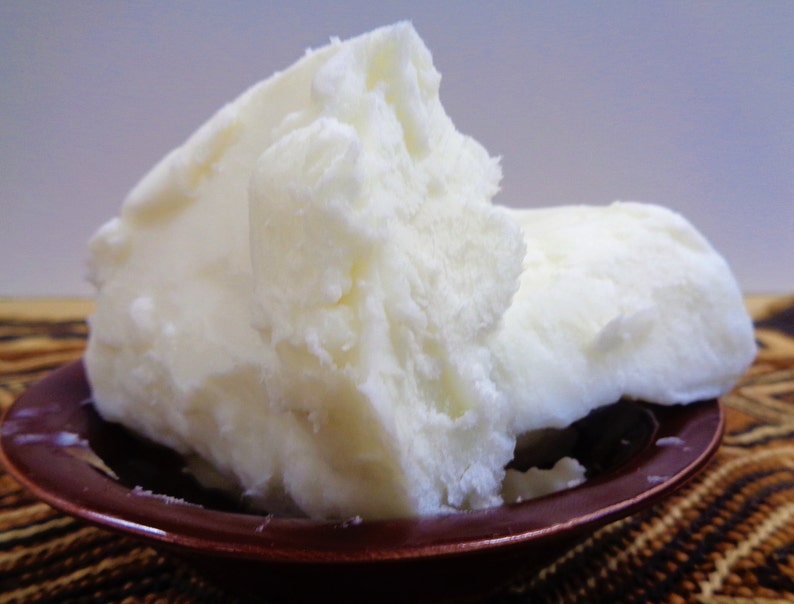 May include: A large, solid, off-white chunk of raw shea butter with a crumbly, uneven texture. The butter is piled high and rests inside a small, round, dark brown bowl with a glossy finish, showcasing its dense and opaque physical appearance.