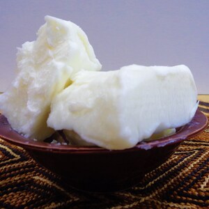 May include: Several large, irregular chunks of solid, opaque white shea butter are piled high inside a small, round, dark brown bowl. The butter has a dense, waxy, and slightly uneven texture with visible ridges and smooth, matte surfaces.