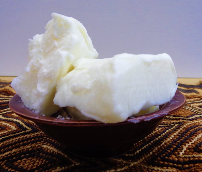 May include: Several large, irregular chunks of solid, opaque white shea butter are piled high inside a small, round, dark brown bowl. The butter has a dense, waxy, and slightly uneven texture with visible ridges and smooth, matte surfaces.
