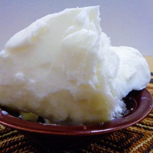 May include: A large, solid, opaque white block of raw shea butter with a crumbly, uneven texture piled high inside a small, round, dark brown bowl. The substance appears dense and waxy, filling the bowl completely with its irregular, jagged edges.