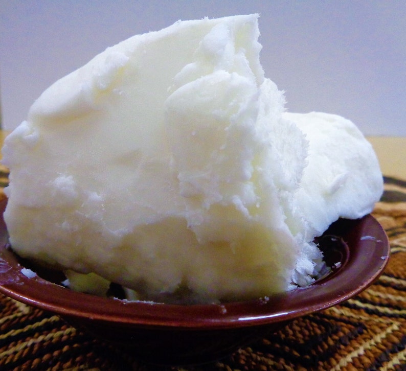 May include: A large, solid, opaque white block of raw shea butter with a crumbly, uneven texture piled high inside a small, round, dark brown bowl. The substance appears dense and waxy, filling the bowl completely with its irregular, jagged edges.
