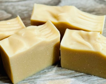 May include: Four bars of handmade soap, each with a light beige color and a smooth, slightly textured surface. The soap bars are arranged on a wooden surface.