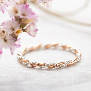 Könnte beinhalten: Ein zarter, gedrehter Roségold-Bandring mit kleinen weißen Edelsteinen, die in die Band eingefasst sind. Der Ring ist auf einer weißen Holzoberfläche mit getrockneten rosa Blüten im Hintergrund fotografiert.