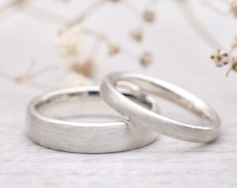 May include: Two silver wedding bands with a brushed finish, lying on a white surface.