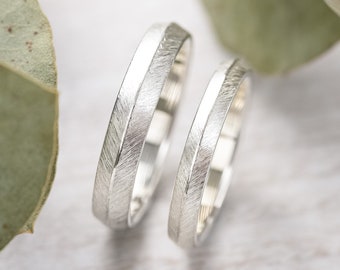 May include: Two silver wedding bands with a brushed finish. The bands are on a white background with green leaves.