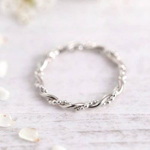 Könnte beinhalten: Ein silberner, gedrehter Ring mit kleinen Perlen auf einer weißen Holzoberfläche mit weißen Blütenblättern, die um ihn herum verstreut sind.