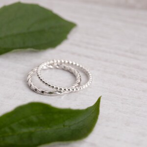 May include: Two silver rings, one with a twisted design and the other with a beaded design, are displayed on a white wooden surface with green leaves.