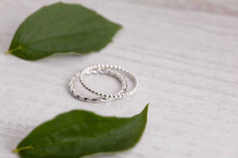 May include: Two silver rings, one with a twisted design and the other with a beaded design, are displayed on a white wooden surface with green leaves.