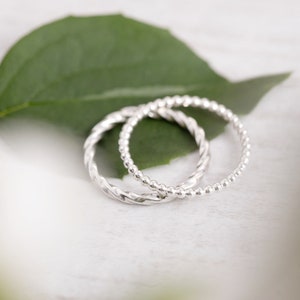 May include: Two silver rings, one with a twisted design and the other with a beaded design, are stacked on top of each other.