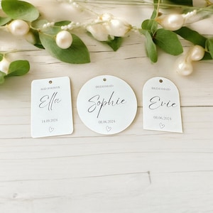 May include: Three white acrylic tags with black text and a small hole at the top. The rectangular tag reads Maid of Honour Ella 14.09.2024, the circular tag reads Bridesmaid Sophie 08.06.2024, and the arched tag reads Bridesmaid Evie 08.06.2024.