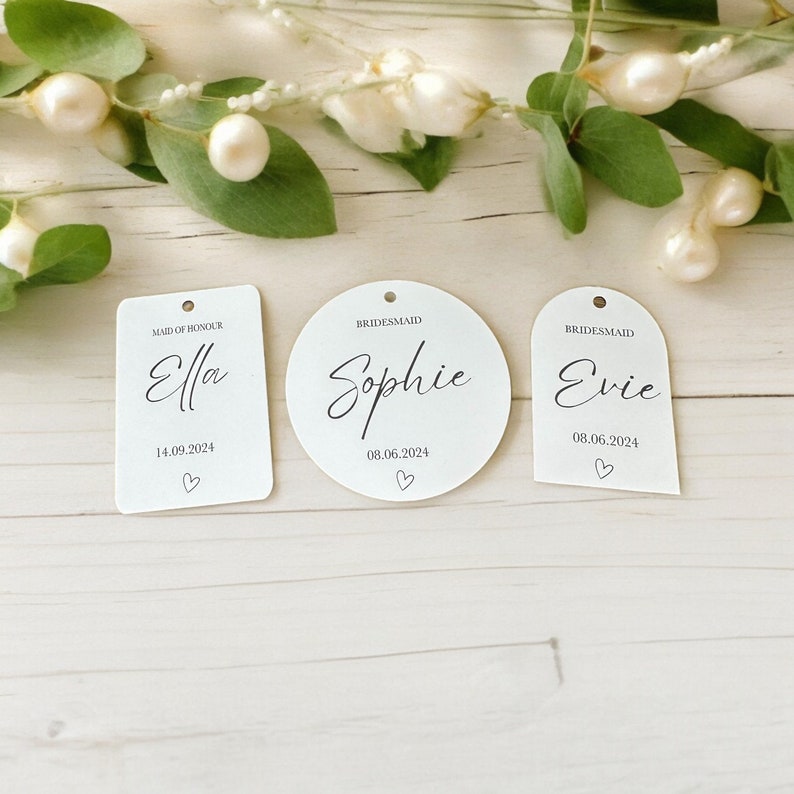 May include: Three white acrylic tags with black text and a small hole at the top. The rectangular tag reads Maid of Honour Ella 14.09.2024, the circular tag reads Bridesmaid Sophie 08.06.2024, and the arched tag reads Bridesmaid Evie 08.06.2024.