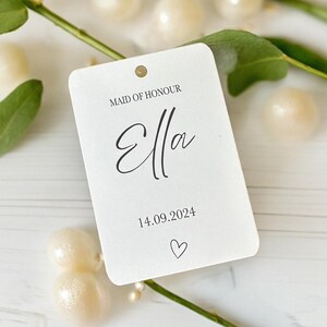 May include: A rectangular white paper tag with rounded corners featuring the black printed text MAID OF HONOUR, the name Ella in a script font, the date 14.09.2024, and a small heart icon. The tag has a single hole punched at the top center.