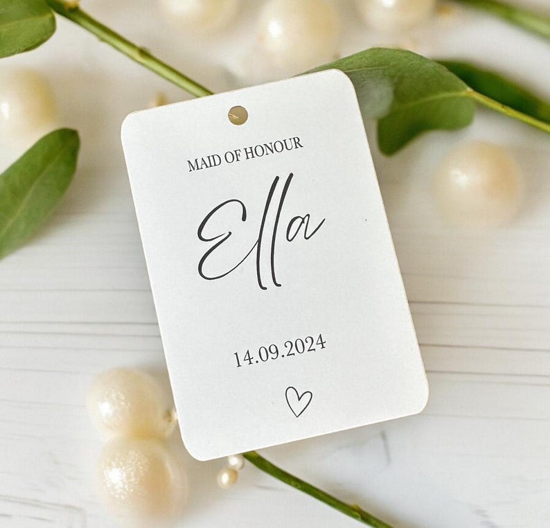 May include: A rectangular white paper tag with rounded corners featuring the black printed text MAID OF HONOUR, the name Ella in a script font, the date 14.09.2024, and a small heart icon. The tag has a single hole punched at the top center.