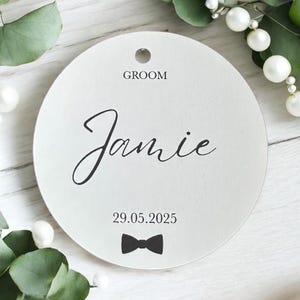 May include: A circular white tag featuring the word GROOM in black text, the name Jamie in a black cursive script, the date 29.05.2025, and a small black bow tie icon. The tag has a hole at the top for hanging and a smooth matte finish.