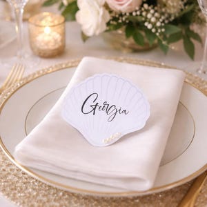 May include: A white seashell place card with the name "Georgia" in script, on a white napkin and gold-rimmed plate. The table setting includes gold cutlery, a gold charger, and a floral arrangement with roses and candles.