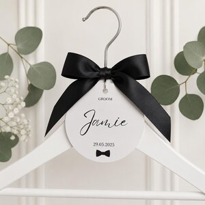 May include: A white wooden clothes hanger featuring a circular white tag with the text GROOM, Jamie, and 29.05.2025, accented by a small black bow tie graphic. A large black satin ribbon is tied in a bow around the silver metal hanger hook.