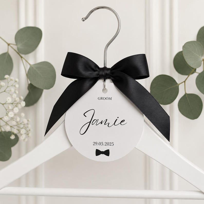 May include: A white wooden clothes hanger featuring a circular white tag with the text GROOM, Jamie, and 29.05.2025, accented by a small black bow tie graphic. A large black satin ribbon is tied in a bow around the silver metal hanger hook.