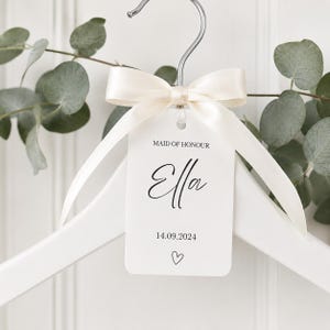 May include: A white wooden clothes hanger featuring a cream-colored satin ribbon tied in a bow around the metal hook. A rectangular white tag hangs from the ribbon, printed with the text MAID OF HONOUR, the name Ella, the date 14.09.2024, and a heart.