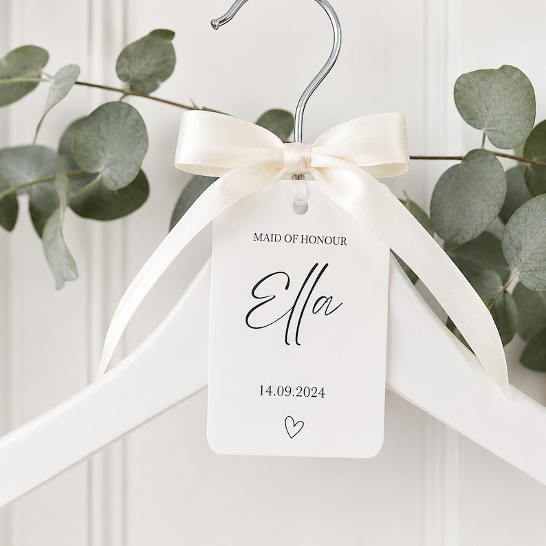 May include: A white wooden clothes hanger featuring a cream-colored satin ribbon tied in a bow around the metal hook. A rectangular white tag hangs from the ribbon, printed with the text MAID OF HONOUR, the name Ella, the date 14.09.2024, and a heart.